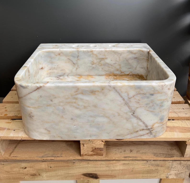 May include: Rectangular marble sink with rounded edges. The sink has a cream and tan colour palette with veining throughout. The sink has three pre-drilled tap holes and is set on a wooden pallet.