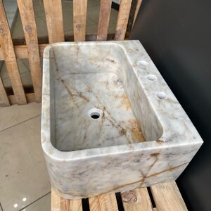 May include: A rectangular marble sink with a rounded front edge. The sink has a central drain hole and three smaller holes on the right side. The marble has a white and beige colour with brown veining. The sink is sitting on a wooden pallet.