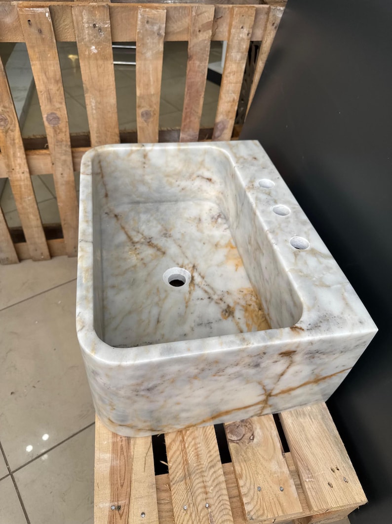 May include: A rectangular marble sink with a rounded front edge. The sink has a central drain hole and three smaller holes on the right side. The marble has a white and beige colour with brown veining. The sink is sitting on a wooden pallet.