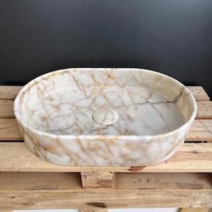 May include: Oval-shaped marble sink with a white and gold veined pattern. The sink has a smooth, polished surface and a central drain. The sink is set on a wooden pallet.