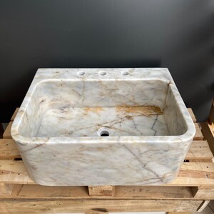 May include: A rectangular marble sink with a light beige and brown veined pattern. The sink has three faucet holes and a single drain hole. It is placed on a wooden pallet.