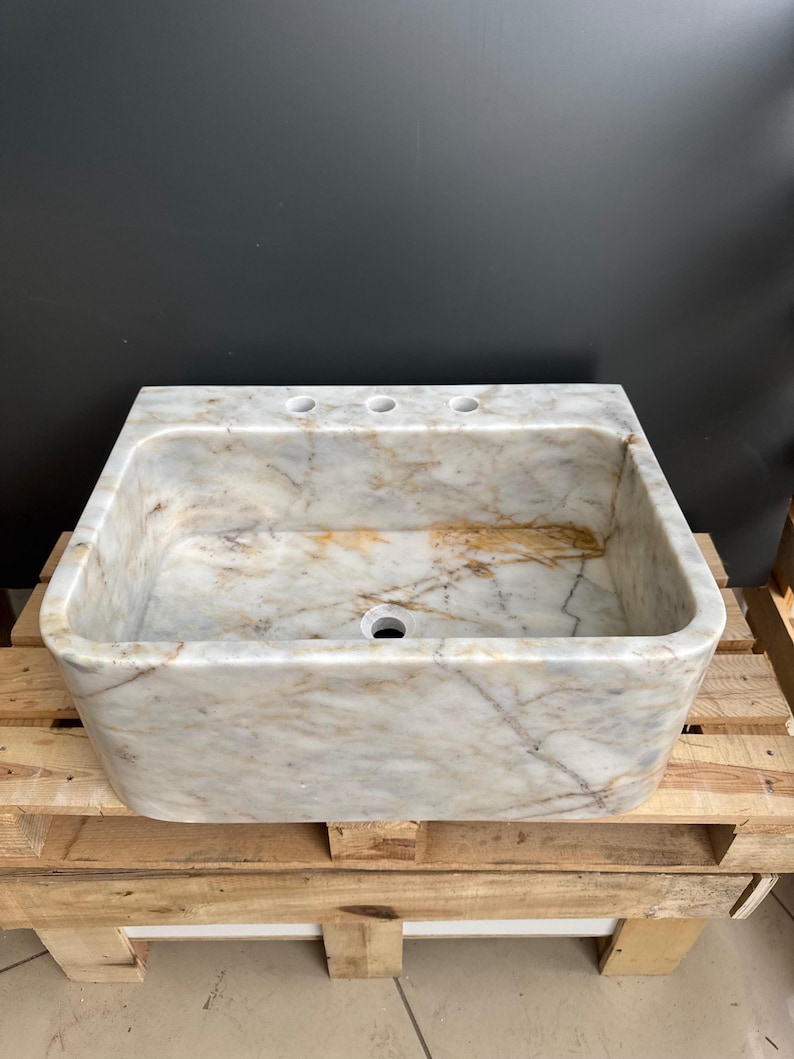 May include: A rectangular marble sink with a light beige and brown veined pattern. The sink has three faucet holes and a single drain hole. It is placed on a wooden pallet.