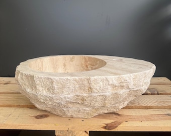 Op de afbeelding: Een beige natuurstenen waskom met een ruwe, getextureerde buitenkant. De kom heeft een cirkelvormig interieur en een kleinere, secundaire opening. De waskom wordt gepresenteerd op een houten pallet.
