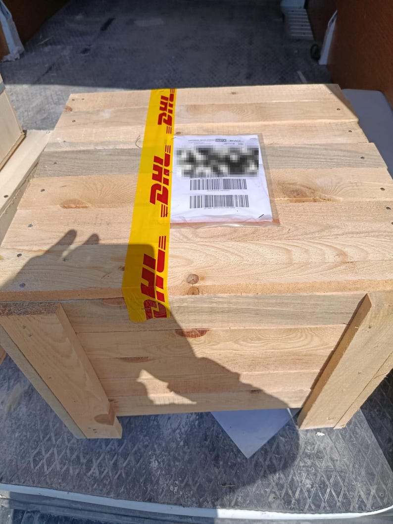 May include: A large, wooden crate constructed from light-colored planks. The crate is secured with yellow DHL tape and has a shipping label attached. The crate is inside a vehicle.