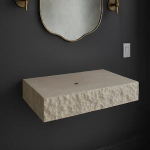 May include: A light beige stone sink with a rough-hewn edge, mounted on a dark grey wall. The rectangular sink has a single drain hole. A gold-framed mirror and sconces are above the sink.