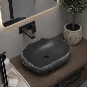 Pode incluir: Uma moderna pia de banheiro feita de pedra cinza escuro com uma borda natural áspera. A pia está sobre uma bancada de madeira com uma torneira preta e um espelho decorativo. Uma pequena planta em vaso adiciona um toque de verde.