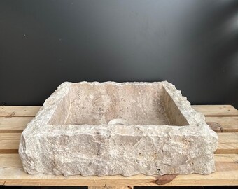 May include: A rectangular, light-colored stone sink with a rough, textured exterior. The sink is set on a wooden pallet, showcasing its natural, rustic aesthetic. The sink's interior is smooth, contrasting with the rugged exterior.