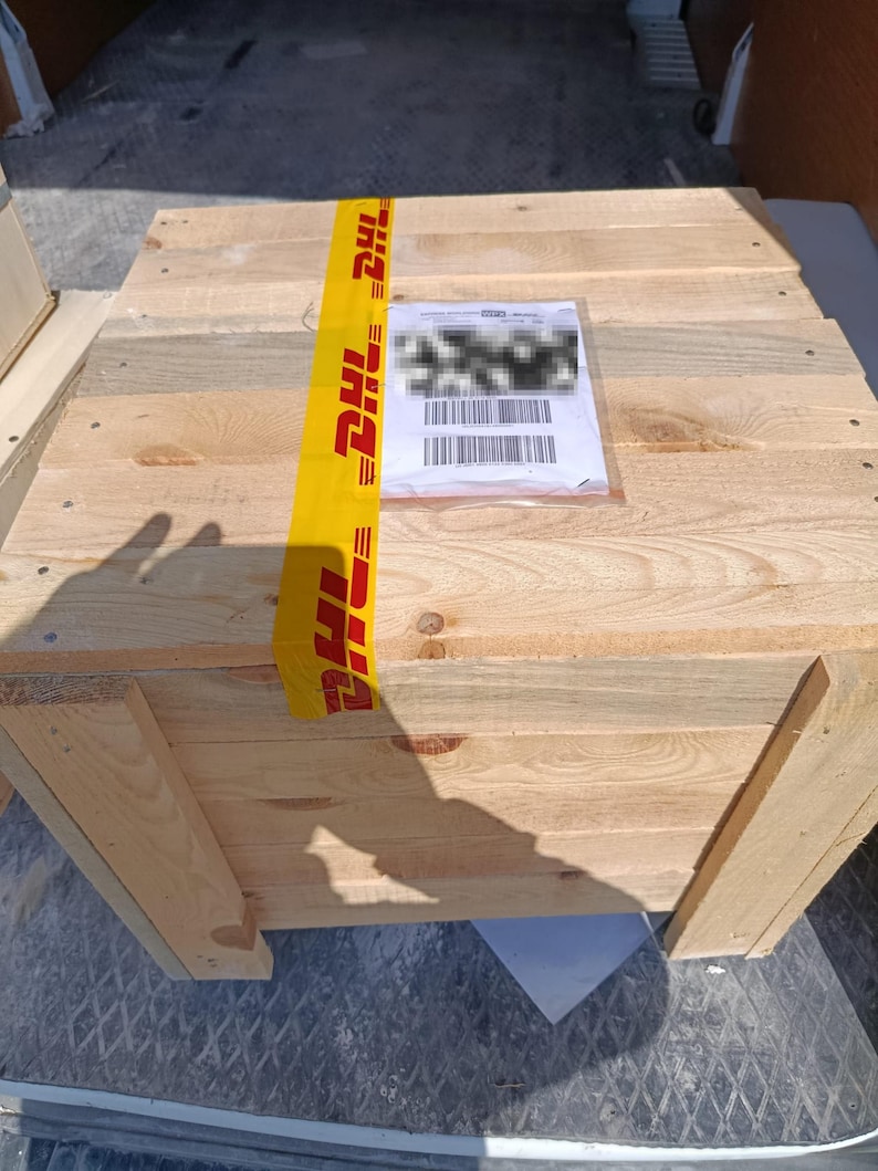 May include: A large wooden crate made from light-coloured planks. The crate is secured with yellow DHL tape and has a shipping label attached. The crate is inside a vehicle.