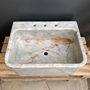 May include: A rectangular marble sink with a light beige and brown veined pattern. The sink has three tap holes and a single drain hole. It is set on a wooden pallet.