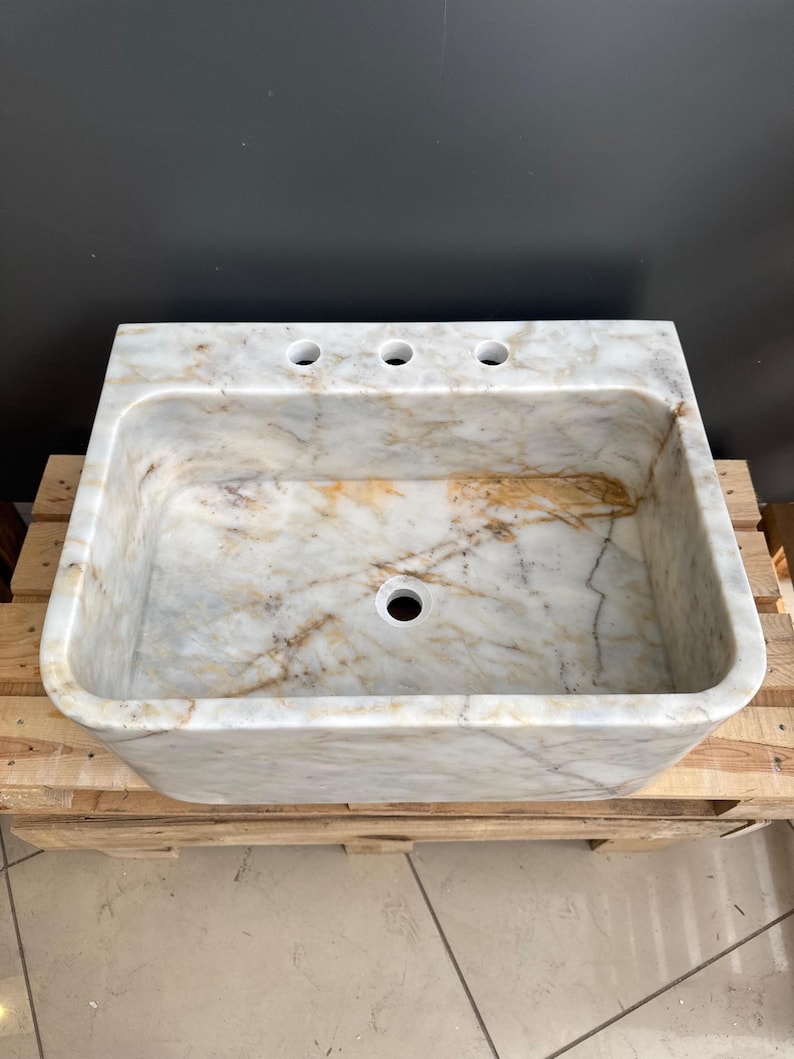 May include: A rectangular marble sink with a light beige and brown veined pattern. The sink has three tap holes and a single drain hole. It is set on a wooden pallet.