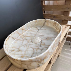 May include: Oval-shaped marble sink with a cream and gold veined pattern. The sink has a smooth, polished finish and a central drain. The sink is made of natural stone and is a bathroom fixture.