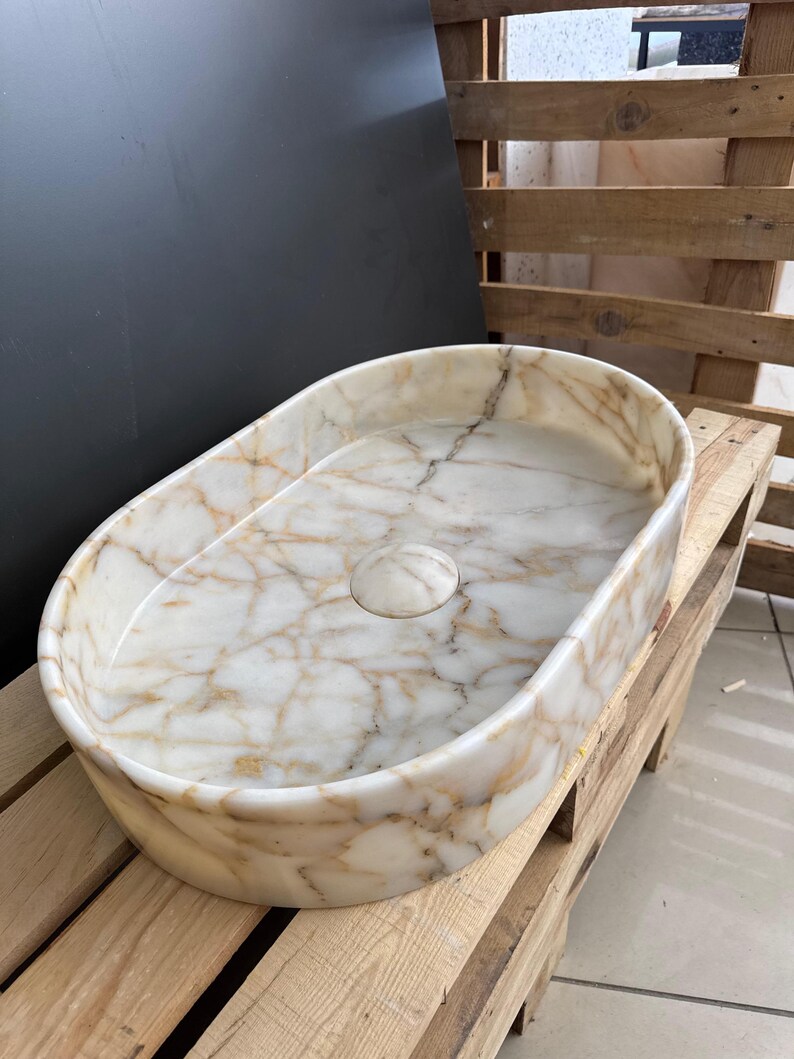 May include: Oval-shaped marble sink with a cream and gold veined pattern. The sink has a smooth, polished finish and a central drain. The sink is made of natural stone and is a bathroom fixture.