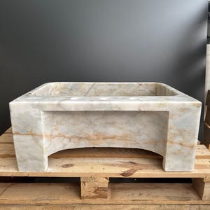 May include: A rectangular marble sink in shades of beige and brown. The sink features a square basin and is supported by a base with arched cutouts. It is presented on a wooden pallet. The sink's dimensions are in centimeters.
