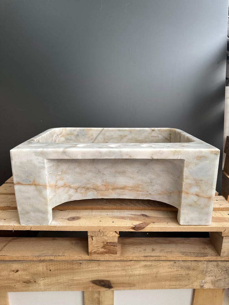 May include: A rectangular marble sink in shades of beige and brown. The sink features a square basin and is supported by a base with arched cutouts. It is presented on a wooden pallet. The sink's dimensions are in centimeters.