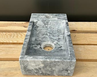 May include: A rectangular, gray marble sink with a rectangular basin and a circular drain. The sink has a natural stone pattern and is set on a wooden pallet.