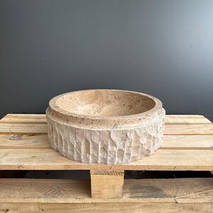 Puede incluir: Un cuenco circular de piedra beige con un exterior texturizado y tallado. El cuenco está sobre un palé de madera. El interior del cuenco es liso, mientras que el exterior tiene un aspecto rugoso y cincelado. El cuenco es un objeto decorativo.