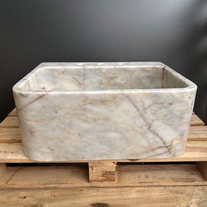 May include: A rectangular marble sink with rounded corners. The sink has a light beige and white colour with brown veining. The sink is sitting on a wooden pallet. The sink is empty.