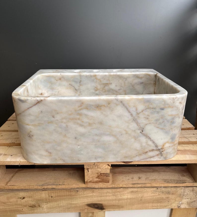 May include: A rectangular marble sink with rounded corners. The sink has a light beige and white colour with brown veining. The sink is sitting on a wooden pallet. The sink is empty.