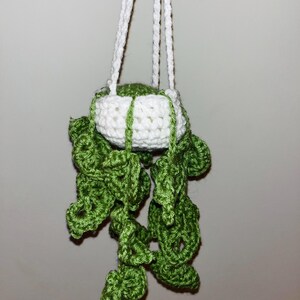 May include: A handmade crochet hanging planter featuring a white pot with green accents and trailing green leaves. The planter is suspended by white cords, creating a charming, handcrafted aesthetic. This item is perfect for adding a touch of nature to any space.