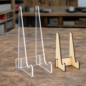 May include: Two display stands, one clear acrylic and one light wood. The acrylic stand has a rectangular base and two angled supports. The wooden stand has a similar design, with a natural wood finish and dark brown edges. Both stands are designed to hold items.