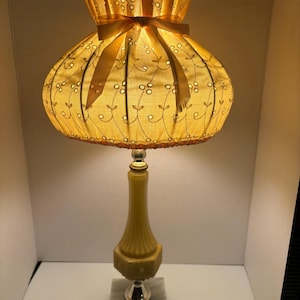 May include: A vintage table lamp with a yellow fabric shade adorned with floral embroidery and a matching ribbon. The lamp has a yellow base with a silver accent and sits on a marble base. The lamp is lit, casting a warm glow.