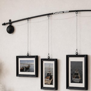 May include: A black curved metal wall-mounted rod with the text Custom text printed on it. Three black rectangular picture frames hang from the rod by thin wires, each displaying a photograph of a boat on water with white matting.