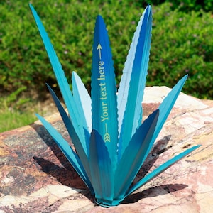 Op de afbeelding: Een metalen agave plant sculptuur in tinten blauw en teal. De bladeren hebben een textuur, geribbeld ontwerp. De tekst "Your text here" is gedrukt op een van de bladeren. De sculptuur wordt getoond op een rots.