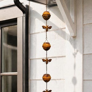 May include: A decorative rain chain with alternating bronze-coloured beehive and bee-shaped ornaments. The chain hangs from a roof overhang, adding a rustic touch to outdoor decor. The beehives are cylindrical with horizontal ridges.