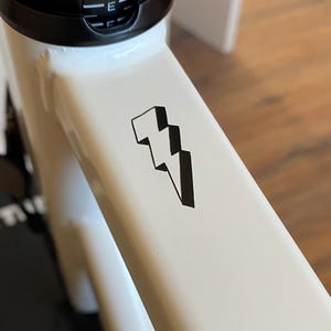 May include: Close-up of a white exercise bike frame with a black lightning bolt graphic. The bike has a black seat post and a water bottle holder. The background includes a black mat and wooden flooring.