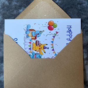 K&ouml;nnte beinhalten: Eine Geburtstagskarte mit Zeichentrickfiguren, darunter ein gelbes Pikachu, ein blaues Schiggy und eine blau-wei&szlig;e Kreatur, die mit Kuchen und Luftballons feiern. Die Karte befindet sich in einem braunen Umschlag, mit dem Wort "Happy" sichtbar.