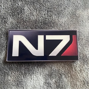 Może przedstawiać: Prostokątna naklejka z białymi literami "N7" na ciemnoniebieskim tle. Logo "N7" ma czerwony akcent po prawej stronie. Naklejka znajduje się na szarej, teksturowanej powierzchni.