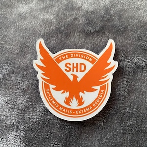 Pode incluir: Um autocolante branco com um emblema de fénix laranja. O autocolante tem o texto "THE DIVISION" no topo, "SHD" no centro e "EXTREMIS MALIS - EXTREMA REMEDIA" na extremidade inferior. O autocolante é circular.
