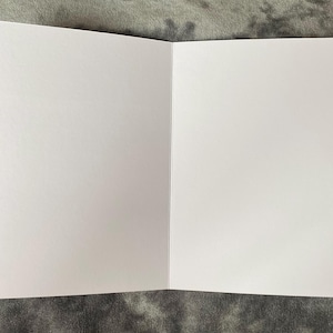 K&ouml;nnte beinhalten: Eine offene, leere Gru&szlig;karte mit wei&szlig;em Innenraum. Die Karte ist aufgeklappt und zeigt zwei leere Seiten. Die Karte liegt vor einem grau strukturierten Hintergrund.