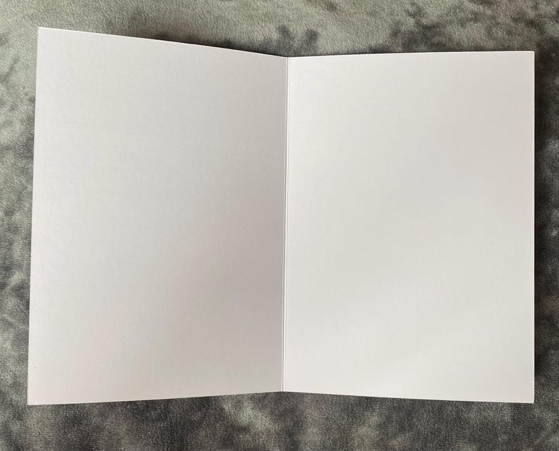 K&ouml;nnte beinhalten: Eine offene, leere Gru&szlig;karte mit wei&szlig;em Innenraum. Die Karte ist aufgeklappt und zeigt zwei leere Seiten. Die Karte liegt vor einem grau strukturierten Hintergrund.