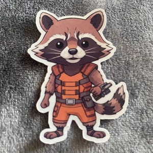 May include: A sticker of a cartoon raccoon character in an orange jumpsuit with a utility belt and a gun. The raccoon has brown fur, a black mask, and a fluffy tail. The sticker has a white border.