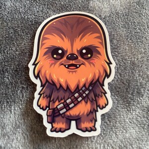 May include: A sticker of a cartoon Chewbacca illustration from Star Wars. The character has brown fur, a bandolier, and a smiling face. The sticker has a white border and is set against a grey background.