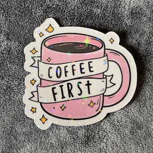 May include: A pink sticker depicting a coffee mug with the text "COFFEE FIRST" on a banner. The mug contains dark coffee and is decorated with yellow stars. The sticker has a holographic finish.