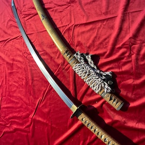 May include: A Japanese katana sword with a polished steel blade and a brown and tan handle. The scabbard is tan with brown accents and a decorative cord. The sword rests on a red velvet surface.