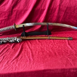 May include: A curved steel katana sword with a wrapped handle resting on a black stand, accompanied by a textured gold-colored scabbard featuring a decorative cord with black and white patterns, displayed horizontally on a red fabric surface.