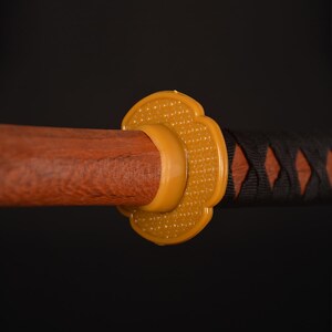 Pu&ograve; includere: Primo piano di una katana. L'impugnatura in legno &egrave; marrone, con una guardia gialla e un'elsa avvolta in nero e marrone. La guardia ha un motivo floreale. La spada &egrave; posta su uno sfondo scuro.