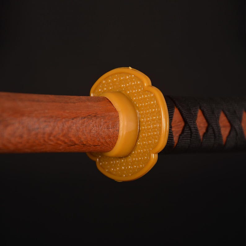 Pu&ograve; includere: Primo piano di una katana. L'impugnatura in legno &egrave; marrone, con una guardia gialla e un'elsa avvolta in nero e marrone. La guardia ha un motivo floreale. La spada &egrave; posta su uno sfondo scuro.