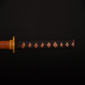 Pu&ograve; includere: Primo piano di un'impugnatura di katana in legno con un rivestimento nero a forma di diamante. L'impugnatura presenta un'impugnatura in legno marrone con intagli a forma di diamante e una guardia color oro. La lama non &egrave; visibile.