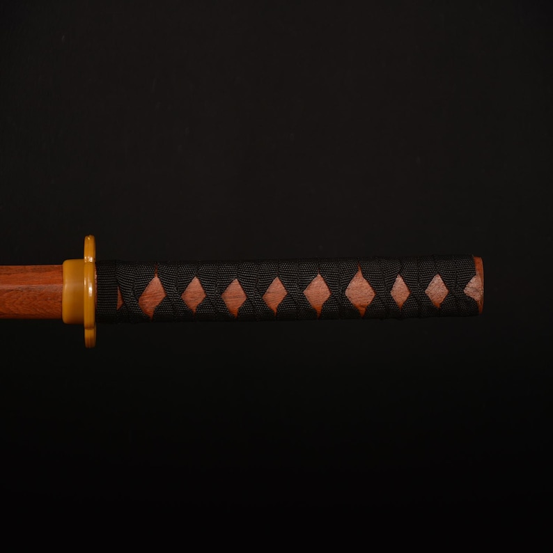 Pu&ograve; includere: Primo piano di un'impugnatura di katana in legno con un rivestimento nero a forma di diamante. L'impugnatura presenta un'impugnatura in legno marrone con intagli a forma di diamante e una guardia color oro. La lama non &egrave; visibile.