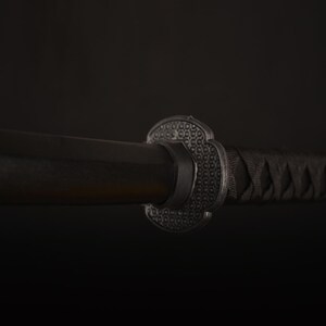 Pu&ograve; includere: Primo piano di una spada katana nera. La spada presenta un'impugnatura in legno scuro, una guardia decorativa con un motivo strutturato e un'impugnatura nera avvolta strettamente. La lama &egrave; parzialmente visibile.