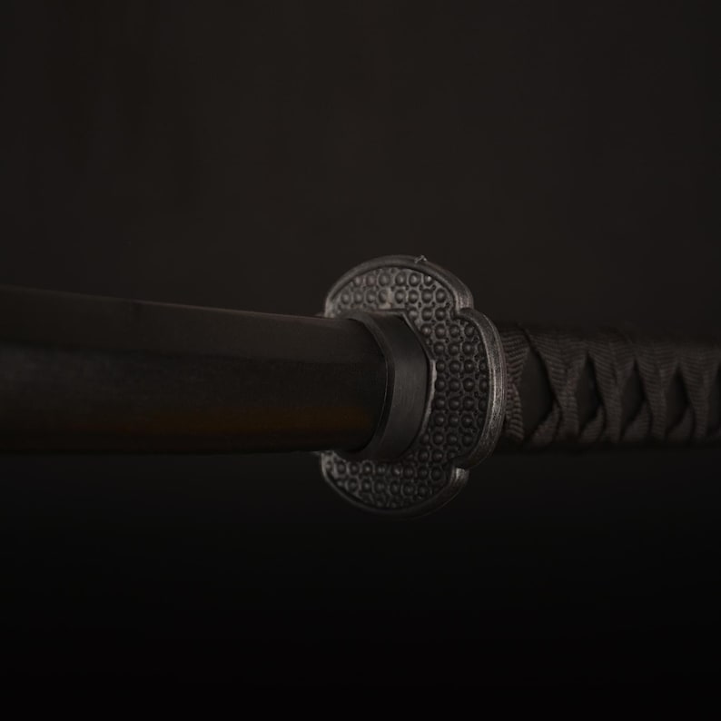 Pu&ograve; includere: Primo piano di una spada katana nera. La spada presenta un'impugnatura in legno scuro, una guardia decorativa con un motivo strutturato e un'impugnatura nera avvolta strettamente. La lama &egrave; parzialmente visibile.