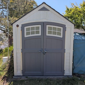 May include: A small, white and grey storage shed with a dark grey door. The shed has two windows with a grid pattern and a small triangular vent at the top. A blue tarpaulin covers an object to the right of the shed.