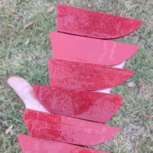 May include: Six red, wedge-shaped wooden pieces are stacked in a hand. The pieces have a glossy finish and are arranged in a fan shape. The background is blurred green grass.