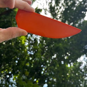 May include: A solid, bright orange, wedge-shaped object with a smooth, matte surface. The item features a curved top edge and a pointed end, resembling a slice of fruit or a geometric segment, held against a natural outdoor background.