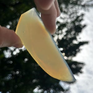 May include: A translucent, wedge-shaped piece of opal gemstone with a smooth, polished surface. The stone displays a pale yellow and milky white coloration, catching the light to reveal subtle internal iridescence and a soft, glowing appearance.