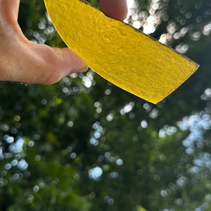 May include: A translucent, bright yellow piece of glass with a curved top edge and a flat bottom edge. The glass has a textured, slightly uneven surface and is held up against the light, showing its vibrant color and semi-transparent quality.